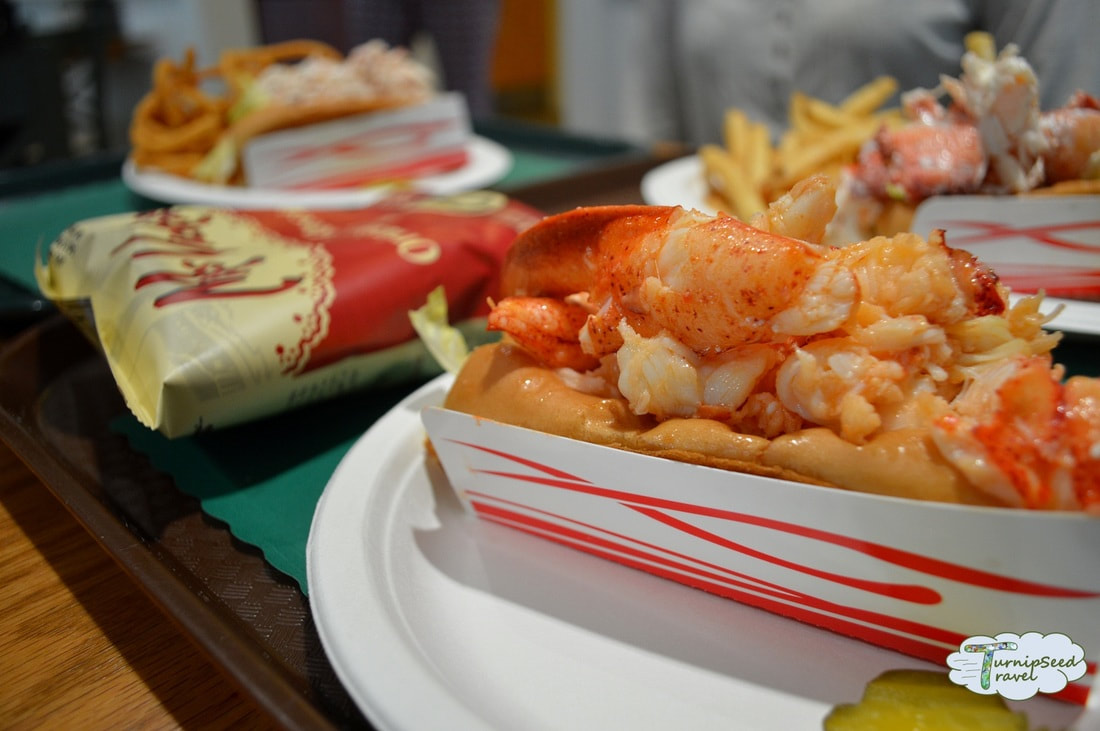 A buttery lobster roll in a red and white cardboard container with other sandwiches and bags of chips in the backgroundture