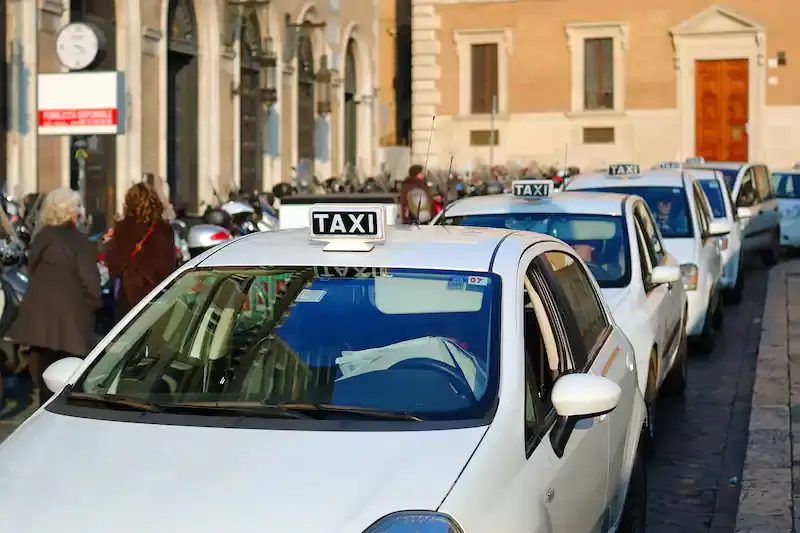 A row of white taxis with people walking and motorcycles parked in the background. Picture
