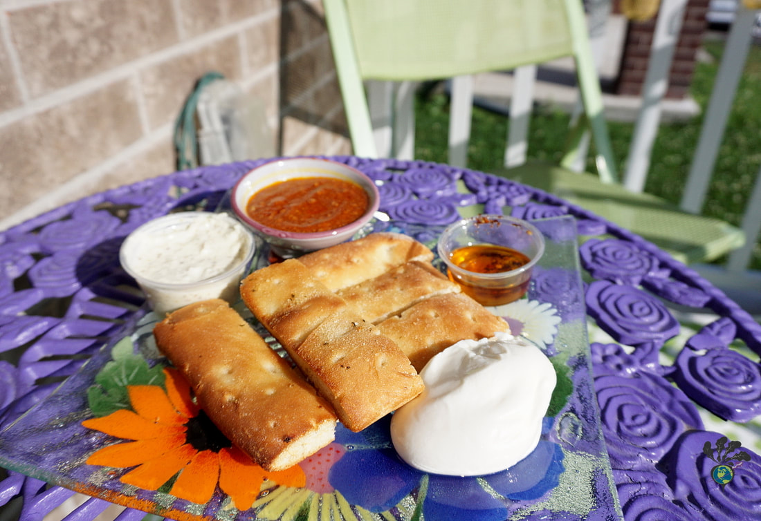 A glass plate with breadsticks, soft cheese, and small containers of dip and tomato sauce sit on a purple patio table 