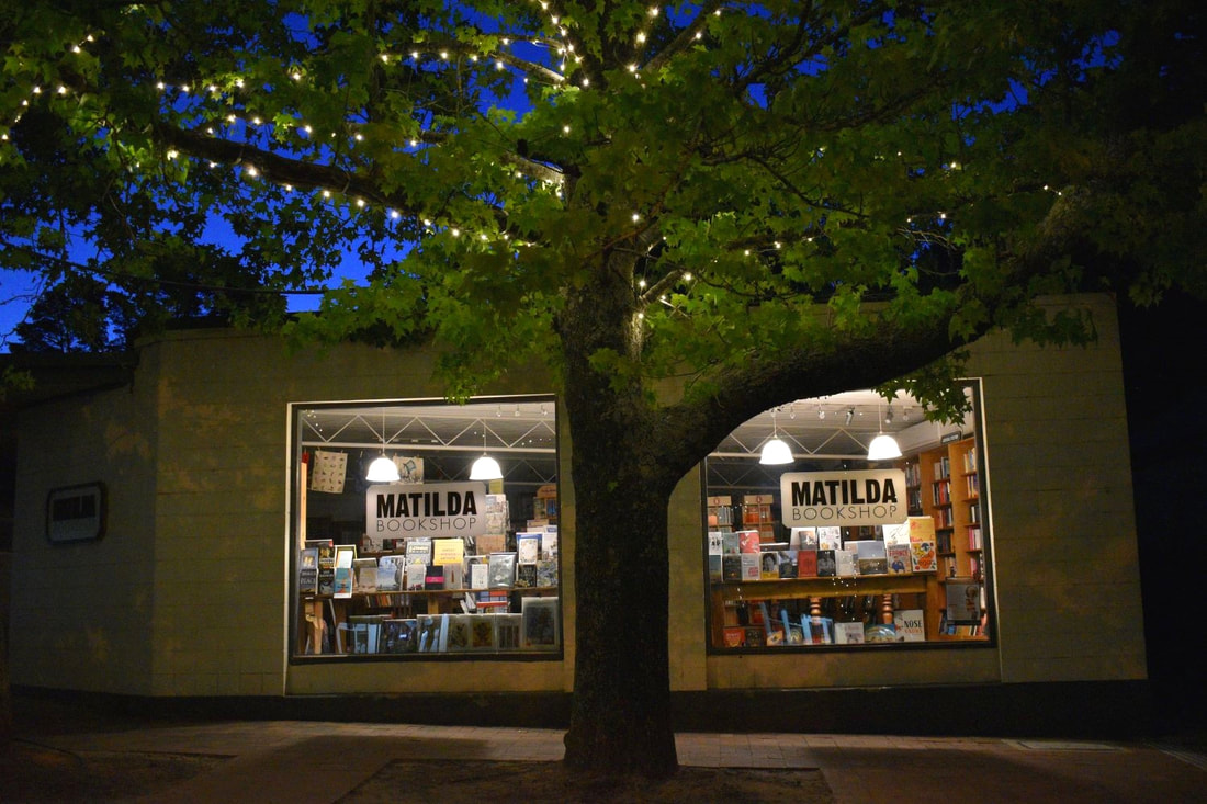 Exterior of the Matilda bookstop at night, with a leafy tree in front of the windows showing bright lights and shelves of books and decorations