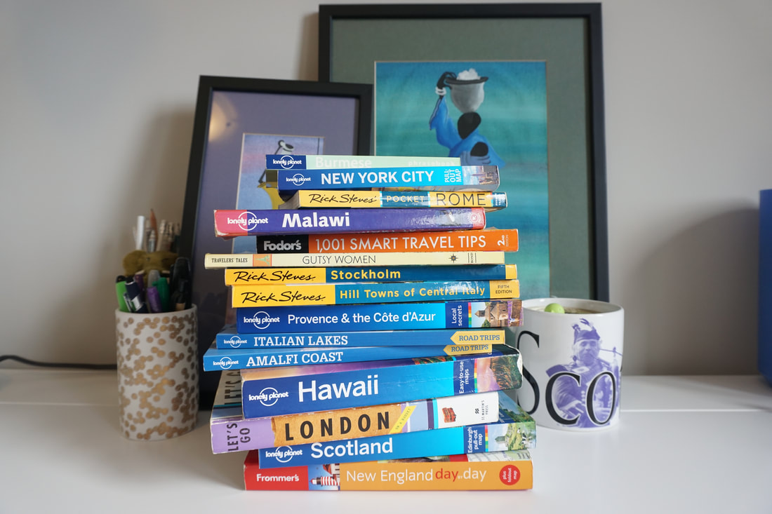 A stack of colorful travel books sit horzontally on top of each other on a white desk with cups of pens and art in the back groundPicture