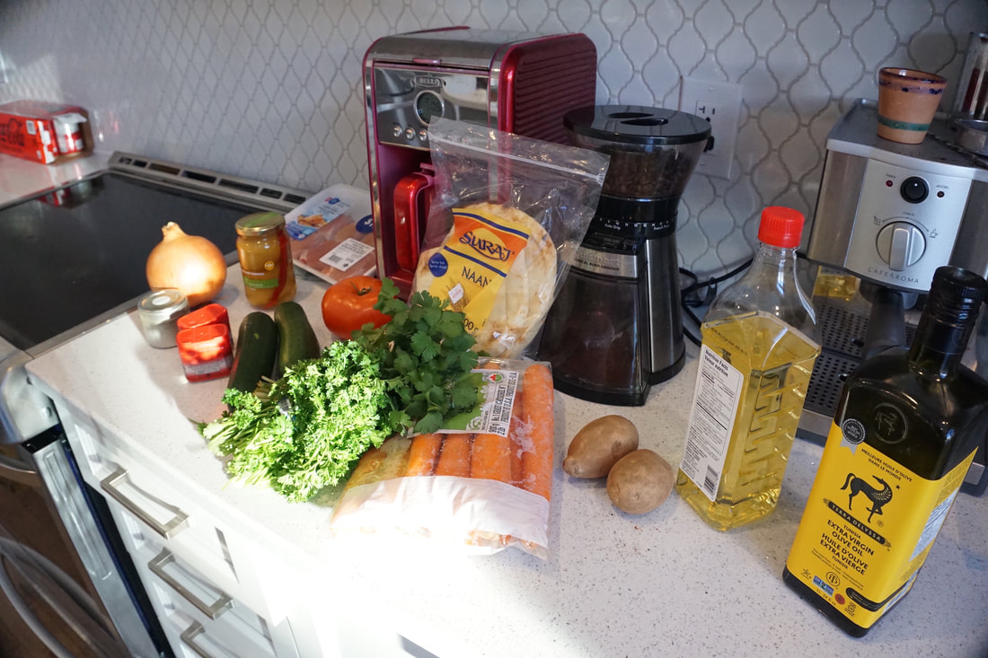 Groceries on the white countertop for the virtual Moroccan cooking class: onion, chicken thighs, containers of spices, herbs, tomato, zucchini, oils.