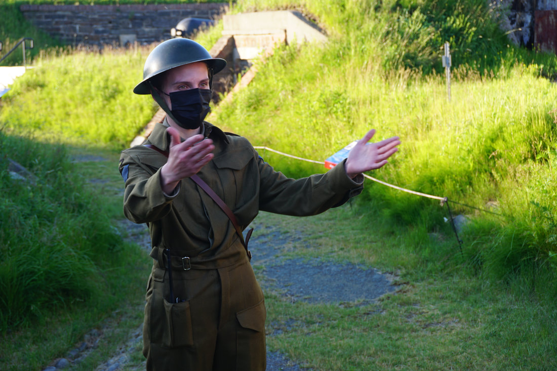 Woman dressed in olive green World War II era uniform and helmut explaining history to a group while standing in front of grassy fortifications.