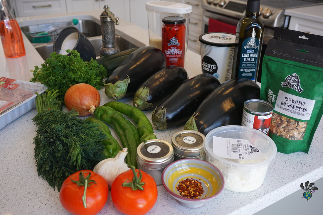 White countertop covered with groceries including several eggplant, peppers, tomatoes, and bunches of herbs, also containers of spices, feta cheese, olive oil, nutsPicture