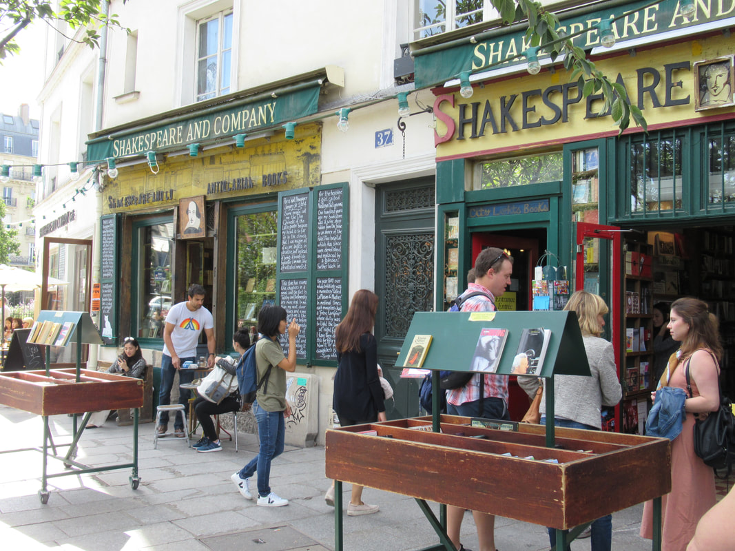 Outside the front doors of Shakespeare and Company bookstore, with green and yellow store front and wooden trays of books out front. Picture