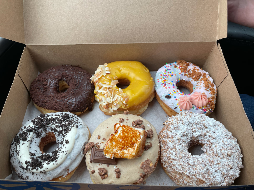 Half a dozen colorful gourmet donuts sit in a cardboard baker's box. Picture