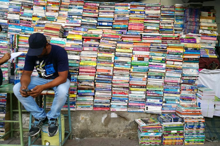 a book seller in India sits on a stool in front a several really high stacks of booksPicture