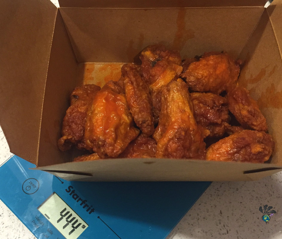 Saucy hot wings sit in a cardboard takeout box on top of a blue scale showing 444 grams