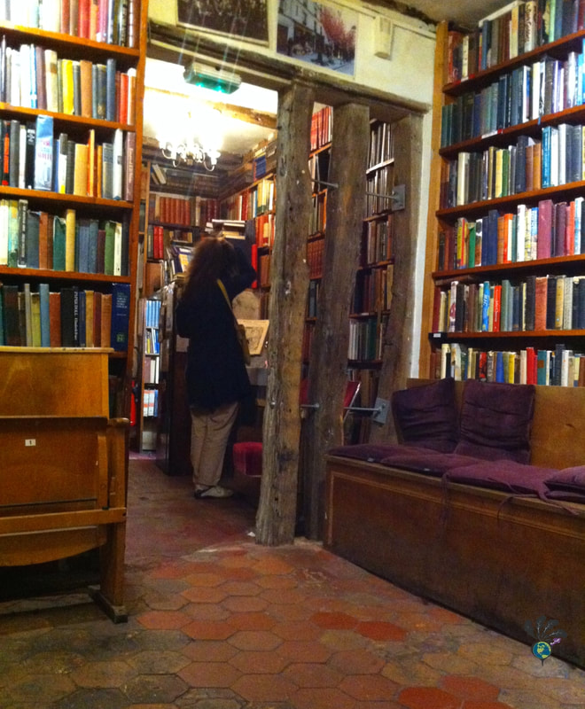 Interior of Shakespeare and Company, showing a woman browsing high bookshelves on a floor of hexagon tilesPicture