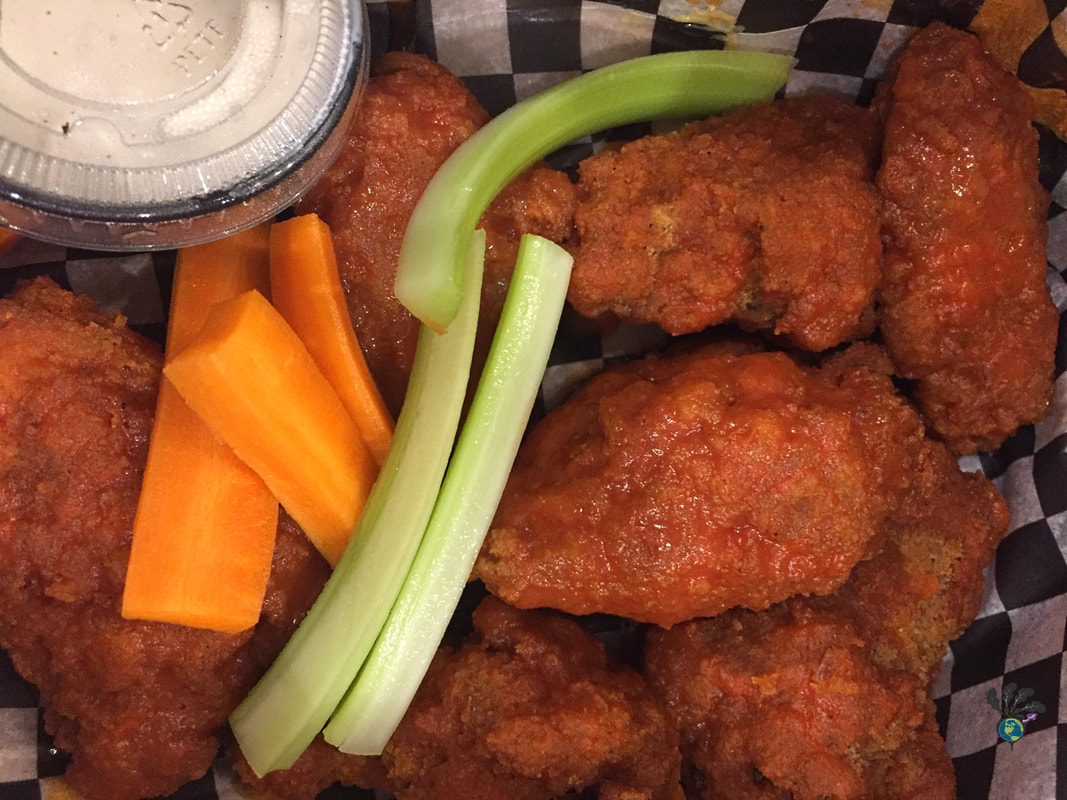 Breaded wings tossed in hot sauce in a takeout container lined with white and black checkered paper, accompanied by a container of ranch sauce and a few carrot and celery sticks.