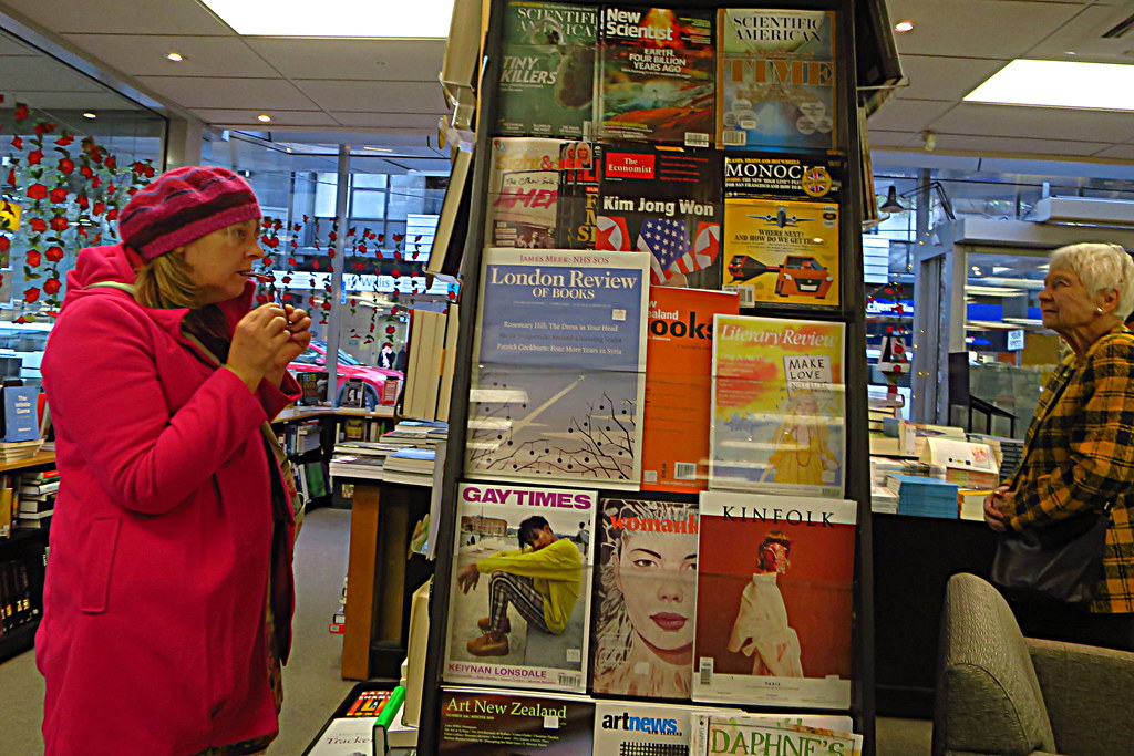 Two women stand on either side of a large colorful magazine rack to look at the selectionPicture