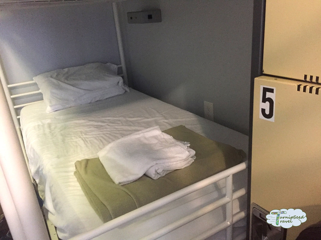 Hostel bunk bed made up with white sheets and a green blanket at the end and a footlocker.Picture