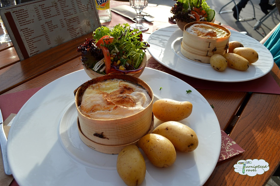 A white plate holds a round of cheese baked in its own rind, four potatoes, and a bowl of salad.