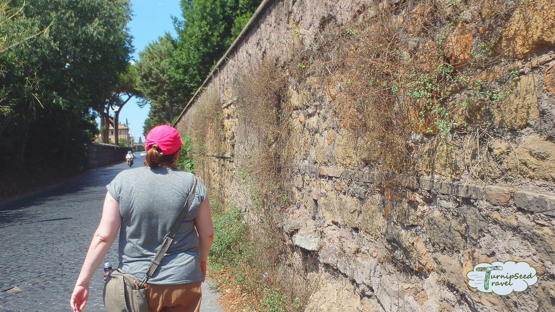 Vanessa walks next to an old stone wall with her back to the camera. You can see a pink welt on her arm next to her grey shirt.Picture