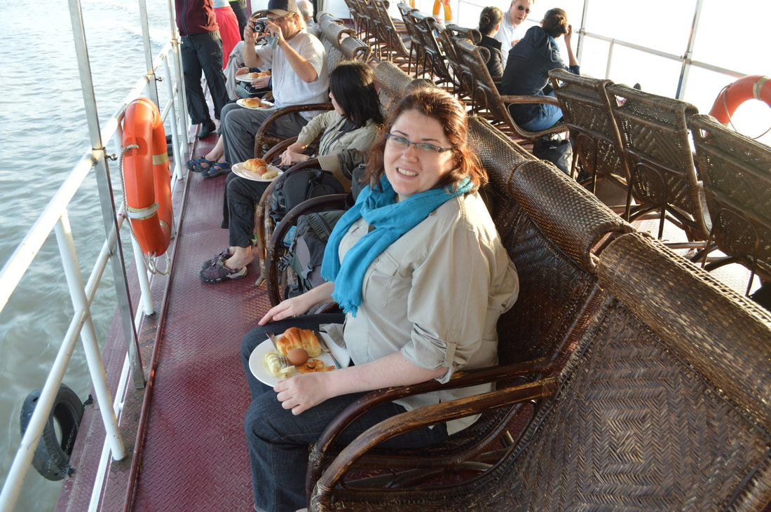 Vanessa sits wearing a white safari shirt and a blue scarf on a rattan deck chair on the board of a ship.