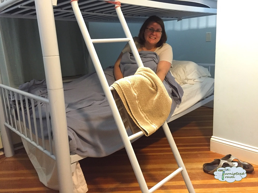 Vanessa sits up in bed in the lower bunk of a white set of bunkbeds in a hostel.