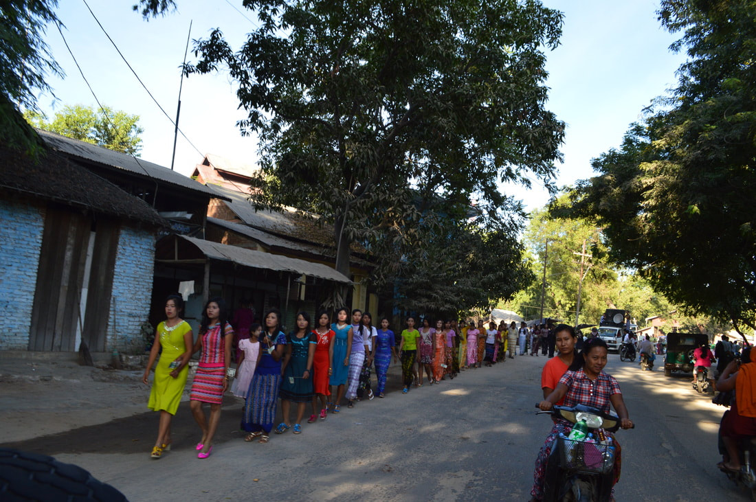 A line of women in colourful dresses walk by the side of the road beside motorcycle and truck traffic