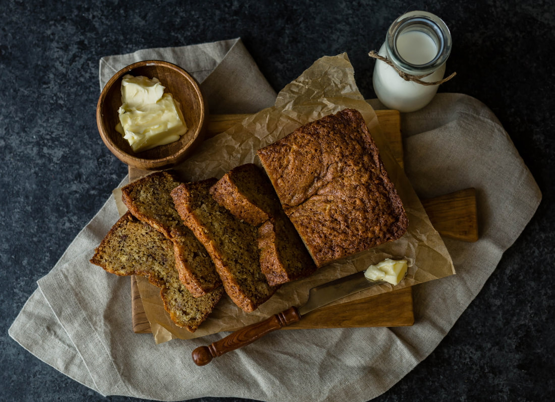 photo of sliced banana bread on waxed paper, wooden cutting board, and linen napkins with bowl of butter and jar of milk in the background