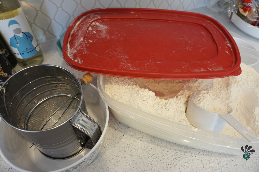A plastic bin of flour with a red lid contains a white measuring cup and a silver sifter sits beside it in a white bowl