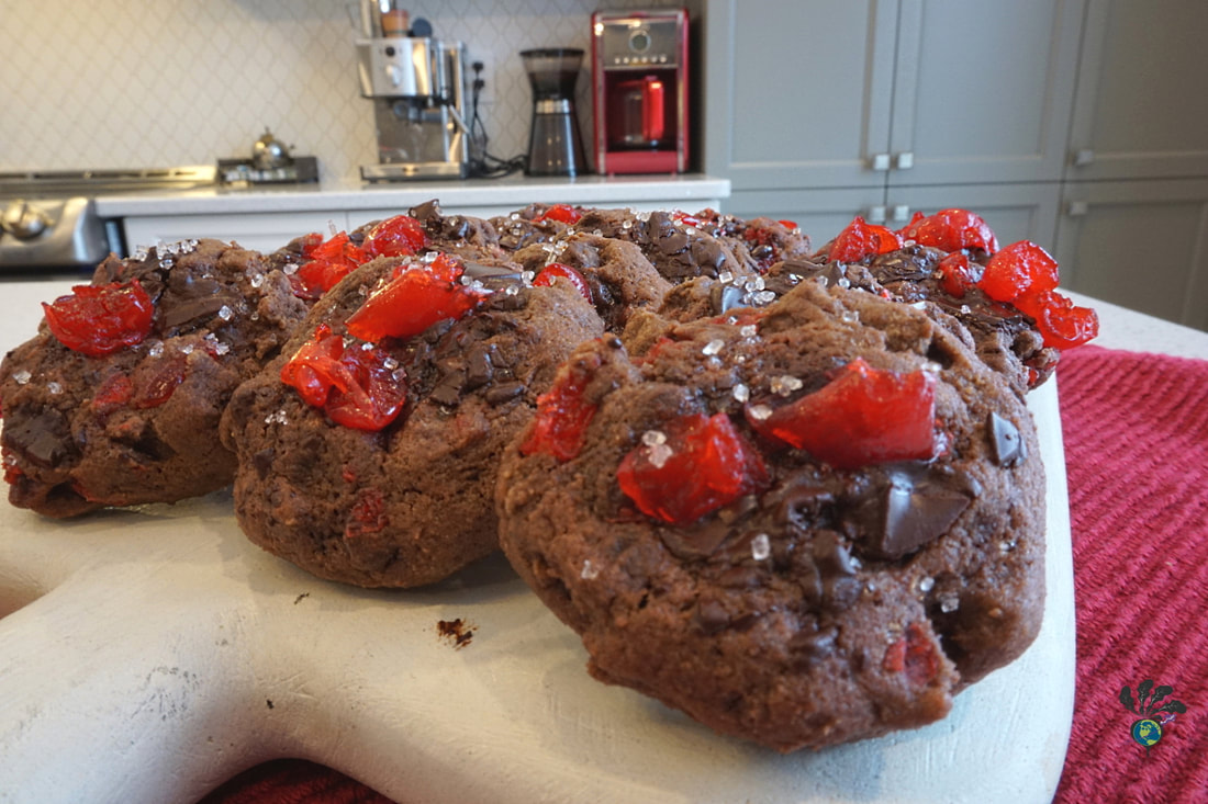 White cutting board on a red cloth, displaying a dozen chocolate cherry cookies with a white kitchen counter in the background.Picture