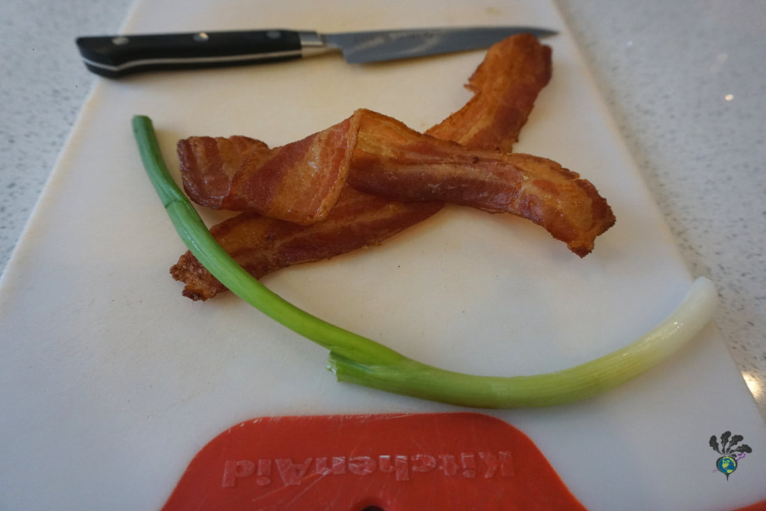 White cutting board with two pieces of bacon, a green onion, and a paring knife 