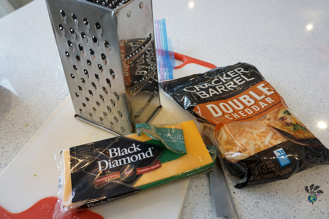 A metal cheese grater sits on a cutting board next to a brick of cheddar cheese and a bag of pre-shredded cheese