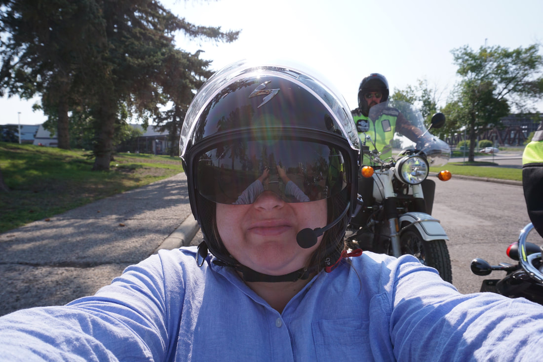 Vanessa takes a selfie while wearing a blue shirt and riding in a motorcycle sidecar, with other riders behind her on a residential street.
