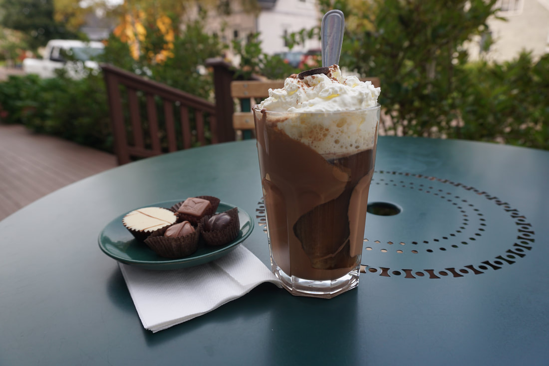 Clear glass filled with chocolate, coffee, and whipped cream, sitting on a green metal outdoor table next to a small plate with chocolate trufflesPicture