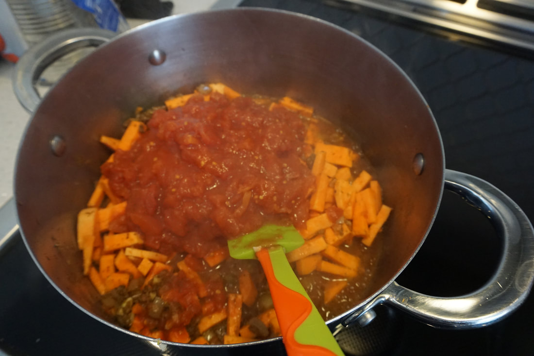 Tomatoes and sweet potatoes are added to the stockpot and stirred with a green spatula 