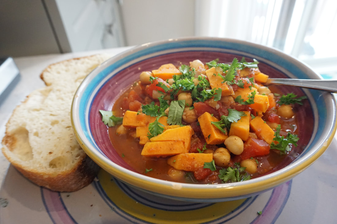 A purple and blue bowl filled with chick pea and sweet potato stew with a spoon in it and a slice of bread on the side.