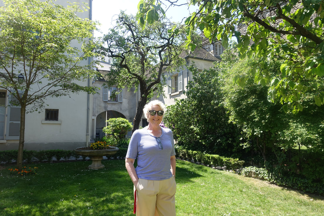 Jo in the garden of the museum with green lawn, shrubs, and white buildings 