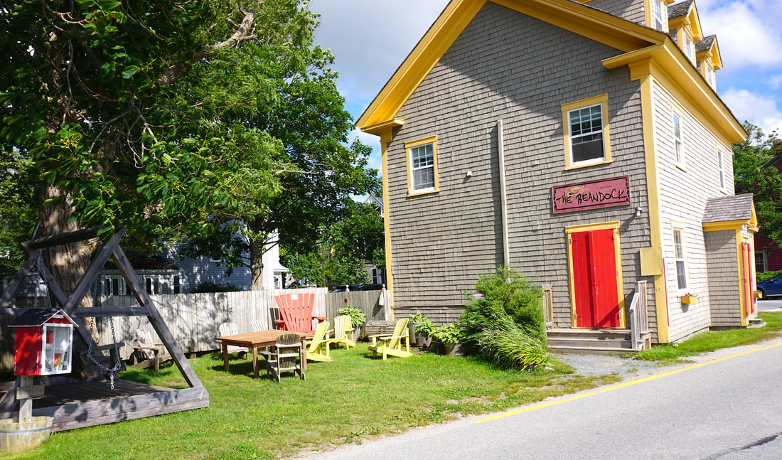 A beige building with bright yellow trim is seen next to a small side yard that holds a red little library box.