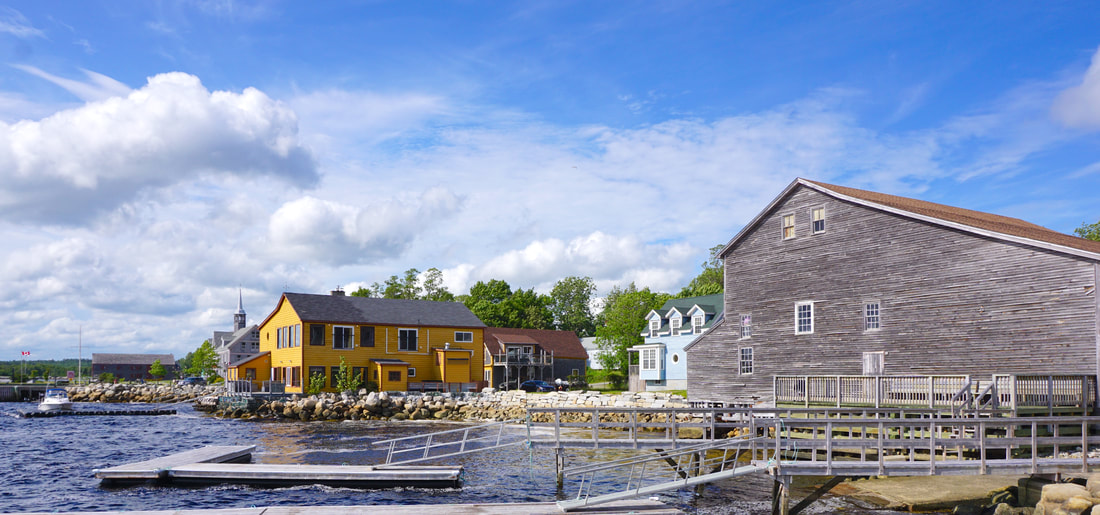 A grey cedar building, a yellow inn, and a church in the distance site at the waterfront, with deep blue water and bright blue skies with fluffy clouds.