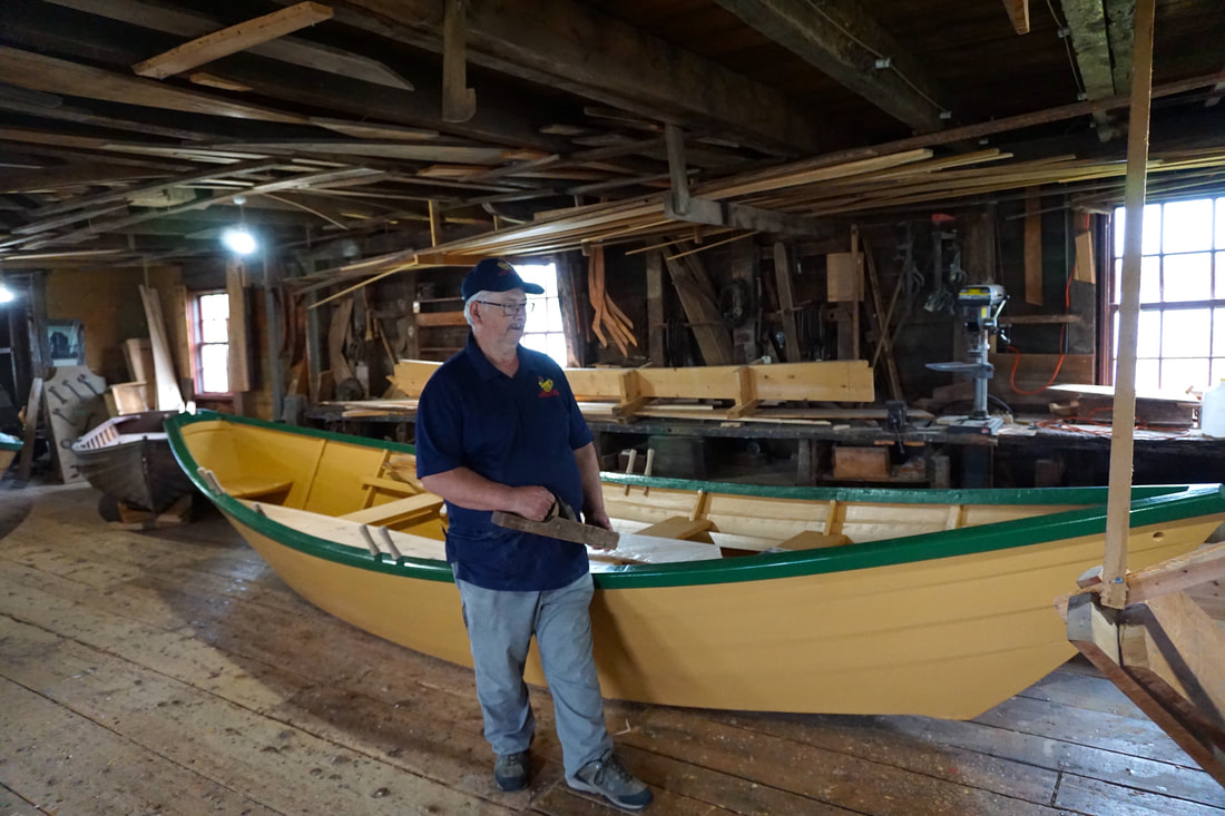 A boat builder, wearing jeans and a blue shirt, stands in front of a pale yellow dory boat with green trim in the workshop