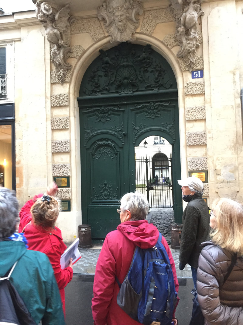 A guide in a red raincoat gestures at a building with an ornate green door while tour participants listen to her.