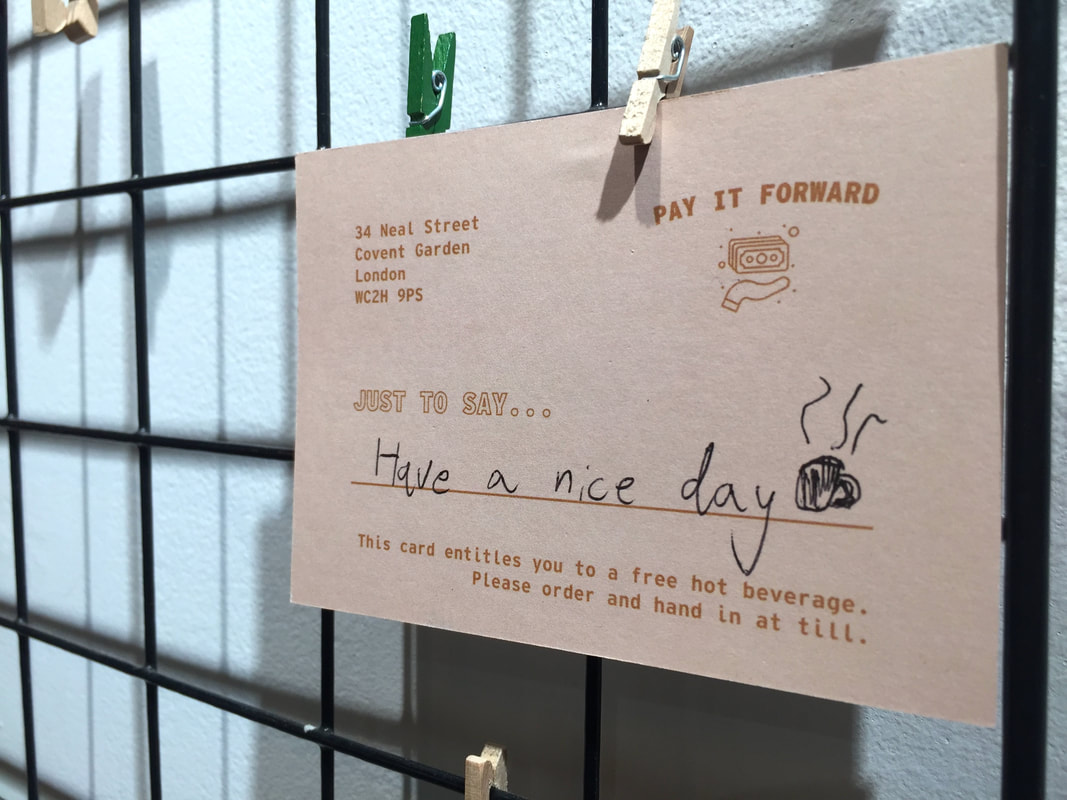 Close up of a cream and brown Pay It Forward Coffee card that has a handwritten message saying 'have a nice day' and a doodle of a coffee mug.Picture