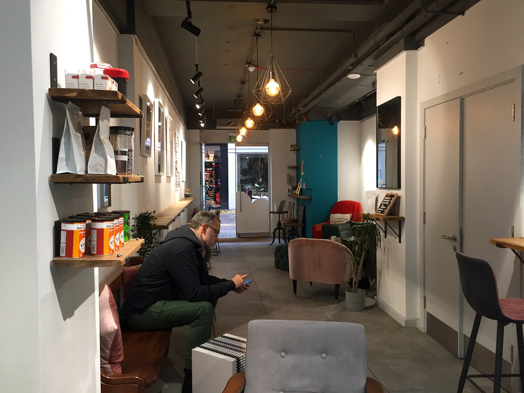 The interior of the shop showing mostly white walls with some teal accents, art on the walls, coffee for sale, and a man in a black coat sitting in a chair and looking at his phone.Picture