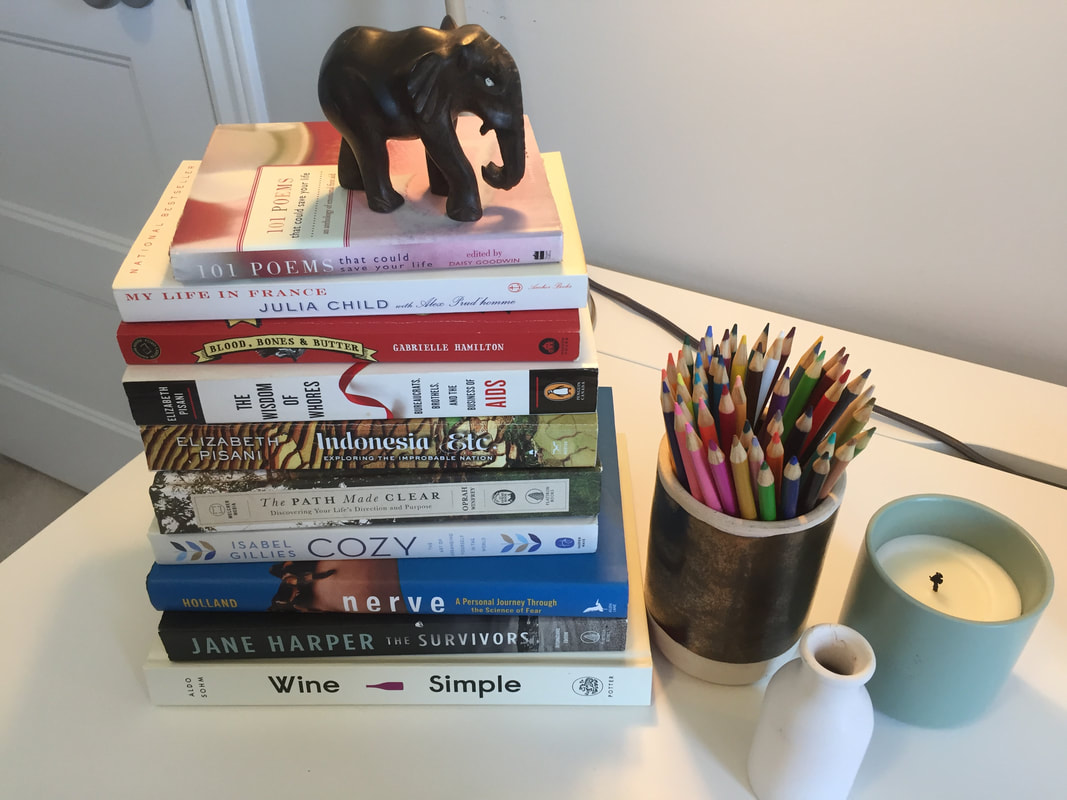 Top view of ten books stacked on top of each other with a small wooden elephant on top, while a container of colourful pencils, a candle, and a small white vase sit beside them.