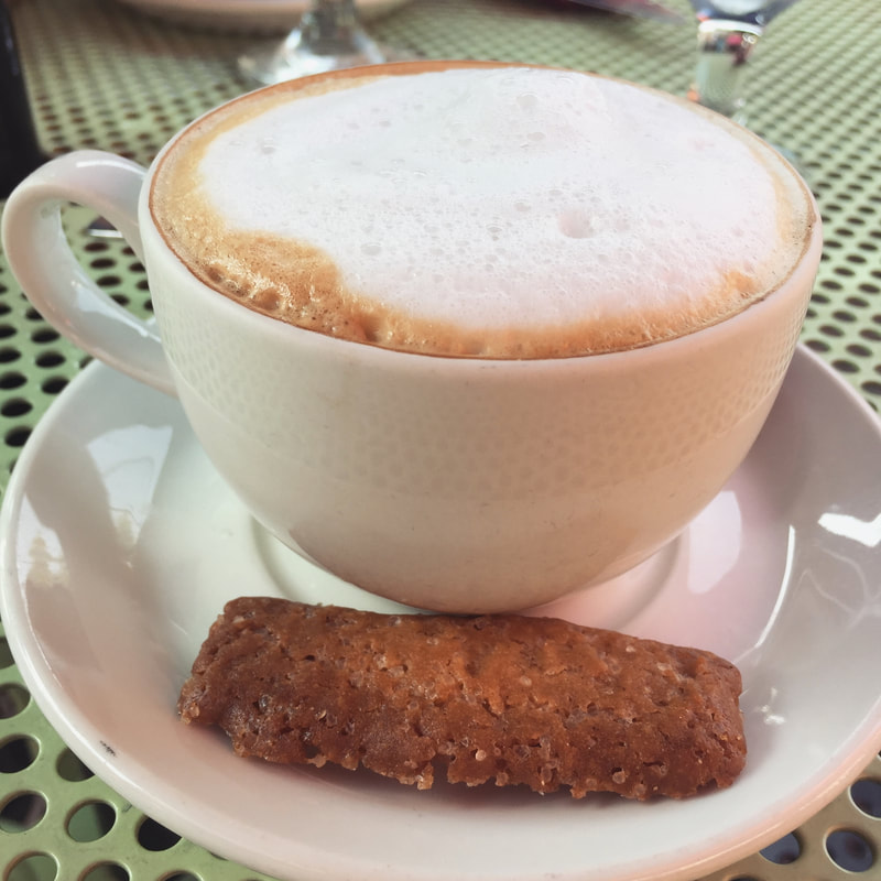 A white mug with foamy cappuchino and a small cookie on a plate