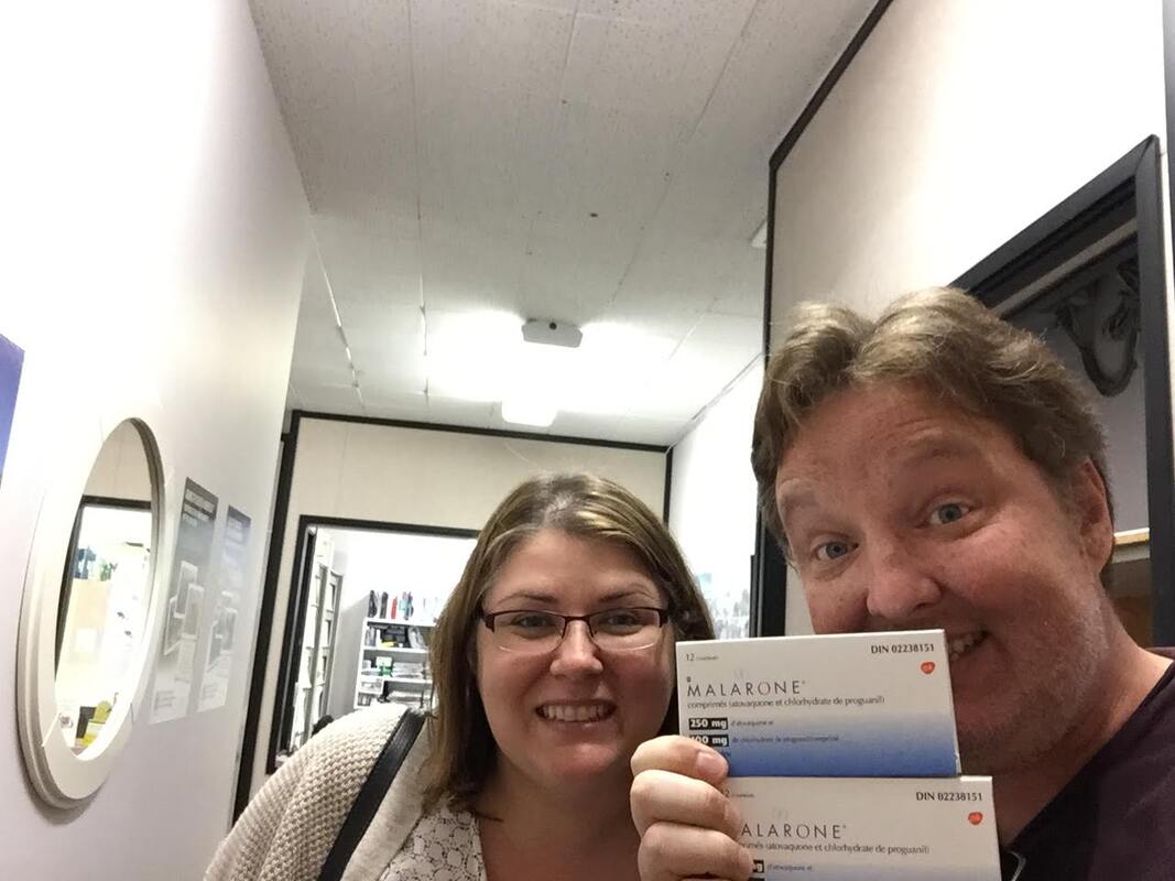 Ryan and Vanessa take a selfie in a travel clinic and hold boxes of medication