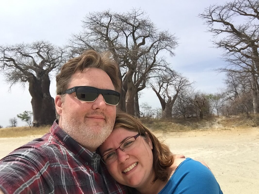 Ryan and Vanessa pose for a selfie with Baobabs in the background surrounded by arid soil.