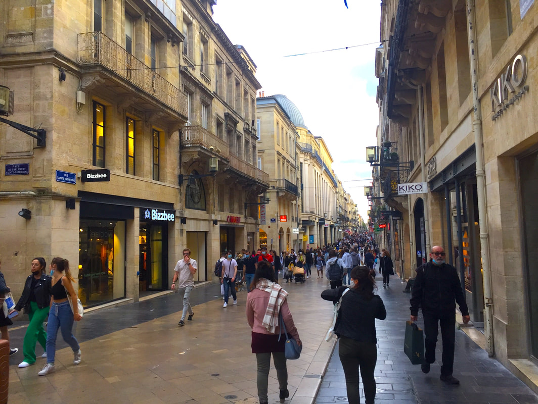Pedestrians walk down a busy shopping street lined with sandstone buildings.