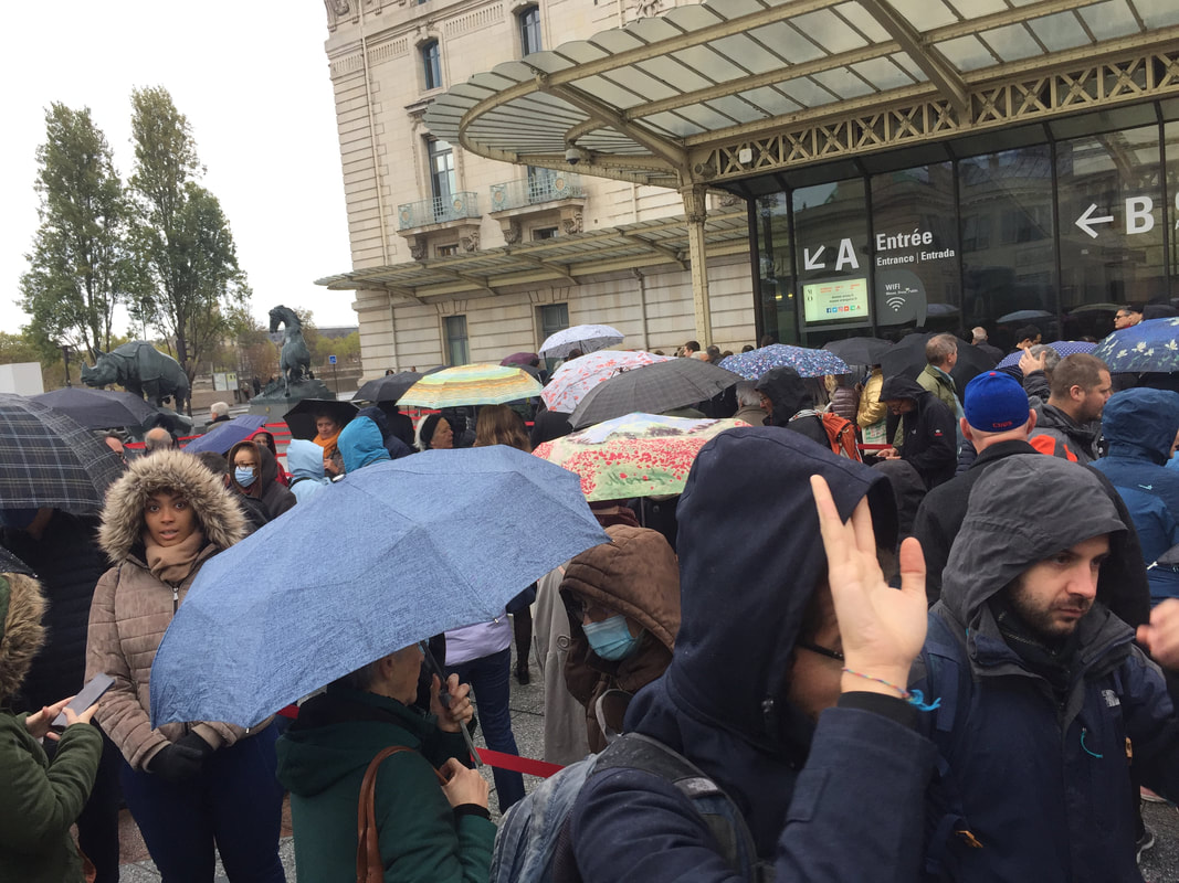 Curving line of people waiting at the Musee D'Orsay, wearing raincoats and holding umbrellas.