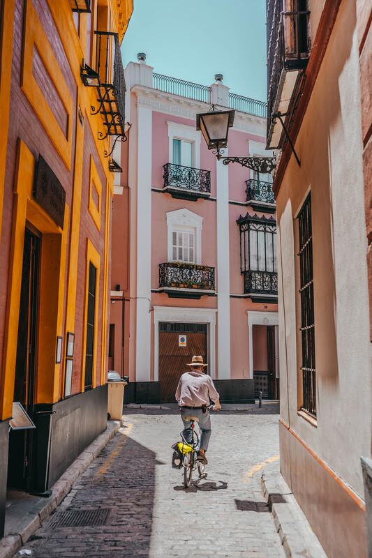 Man ride a bicycle while wearing a sunhat, biking between pink and brown buildings in a narrow laneway