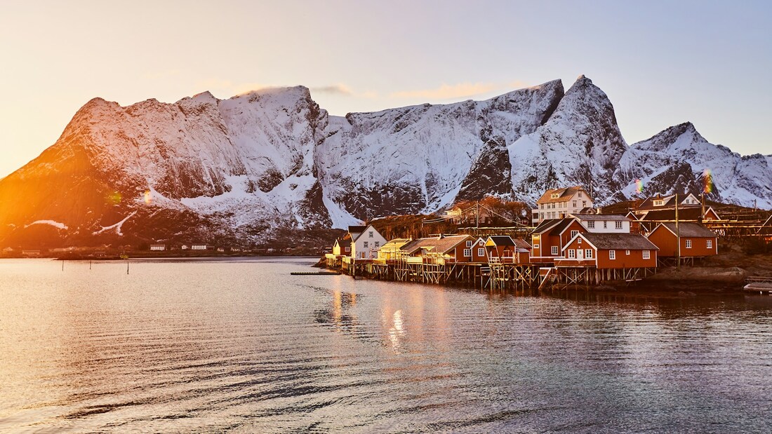 Visit Lofoten Norway: A cluster of houses on stilts at the water's edge with snow covered cliffs in the background.Picture