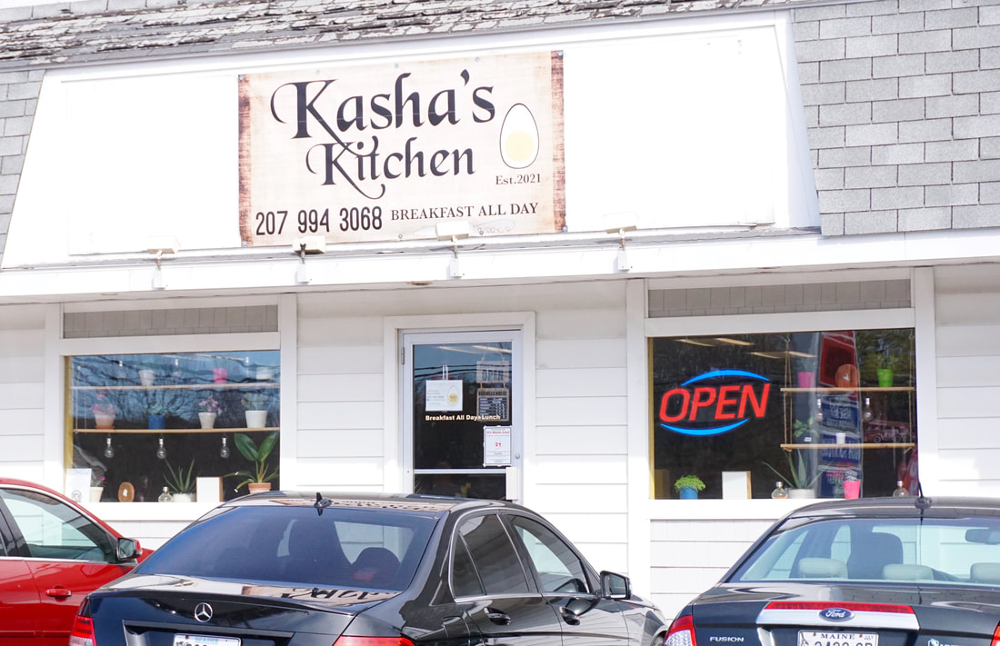 Outside of Kasha's kitchen, showing a white building, large sign, and several cars parked in front.