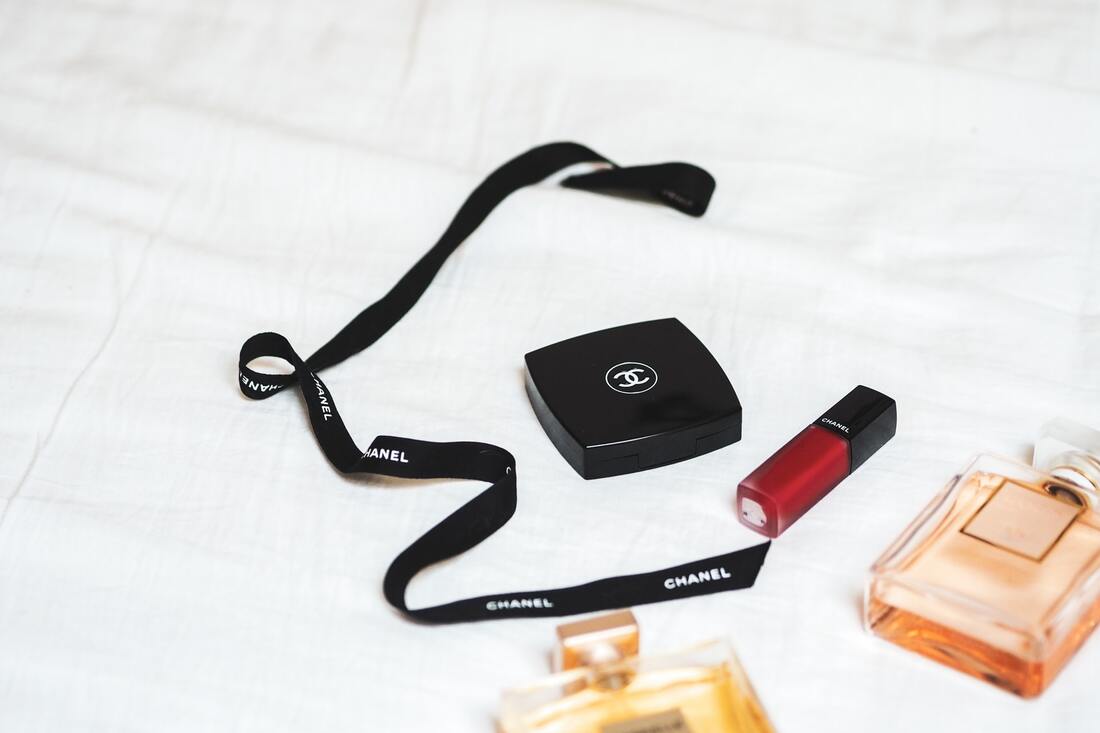Black Chanel ribbon, compact, red lip gloss, and bottle of perfume on white sheet