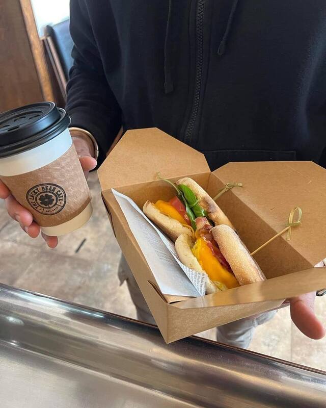 A person holding a to-go coffee cup and a take out box with two breakfast sandwiches on English muffins