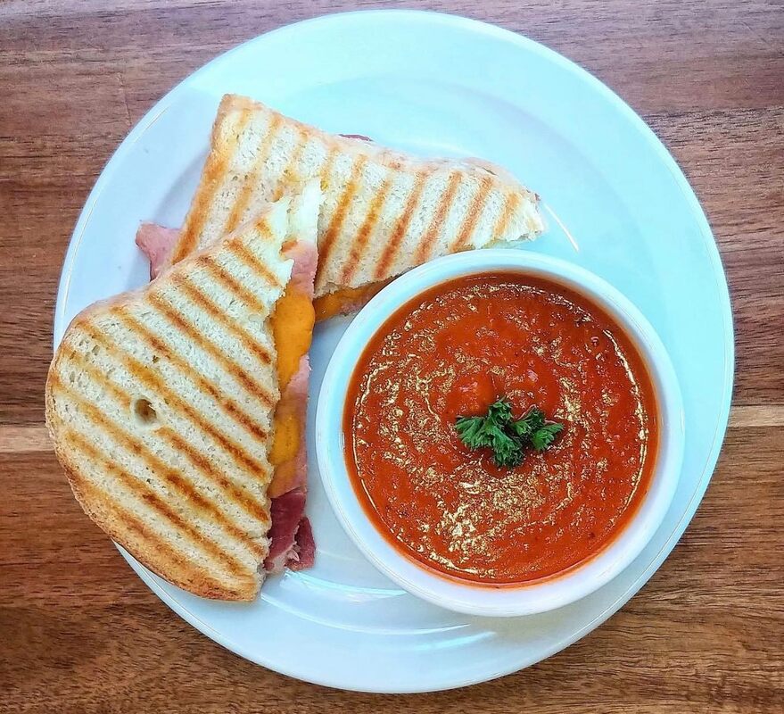 A grilled cheese sandwich with ham and a bowl of tomato soup sit on a white plate.