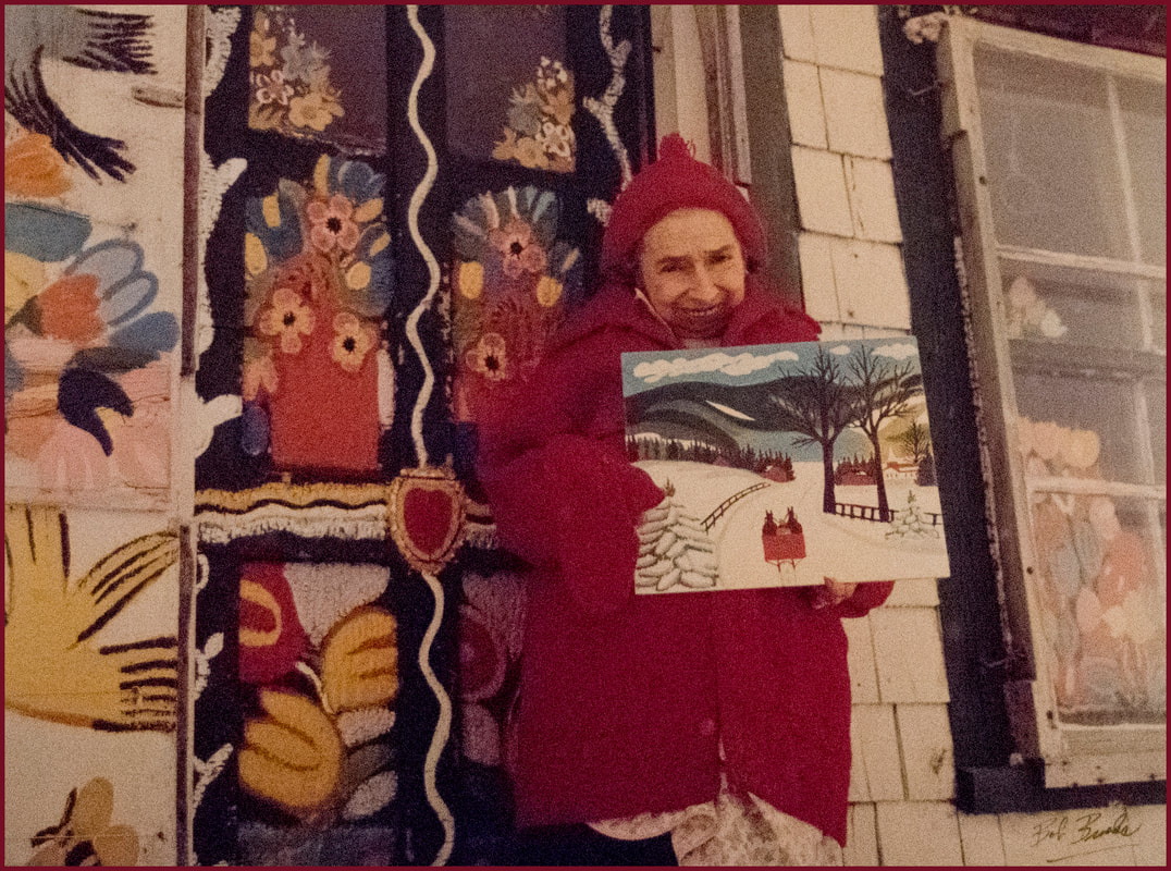 A historic photo of artist Maud Lewis standing outside her home, wearing a red coat, and holding a paintingPicture